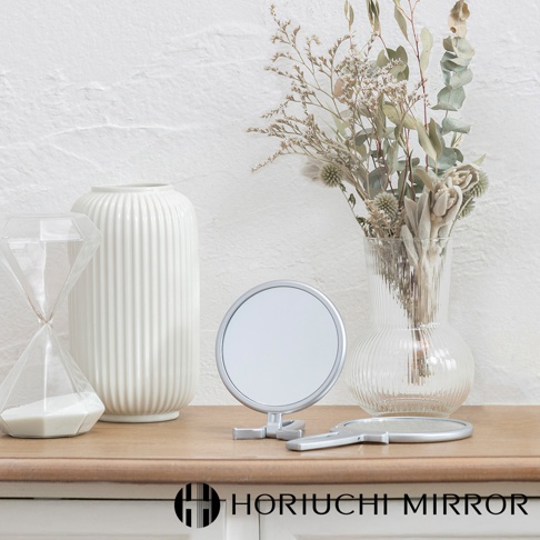 堀內鏡 HORIUCHI MIRROR 的 Napure 手持立式兩用3倍美妝鏡，陳設在木質桌面，背景為白色紋理牆，一旁有玻璃花瓶內的乾燥花束和沙漏裝飾