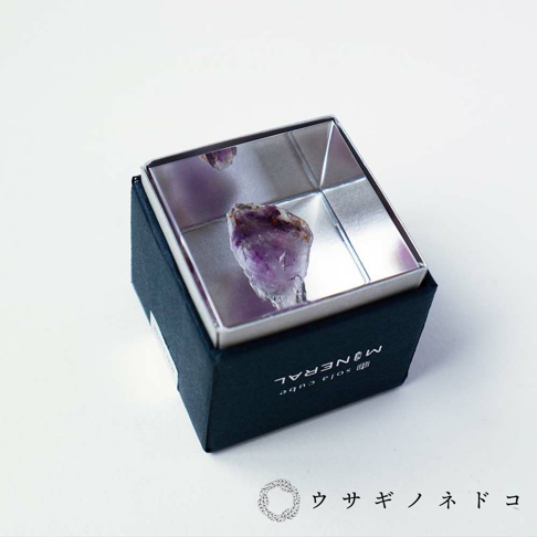 一個黑色盒子內，一個銀色方形展示盒中，放置著一塊天然紫水晶簇，水晶呈現紫色和透明色，表面有明顯的晶體結構。