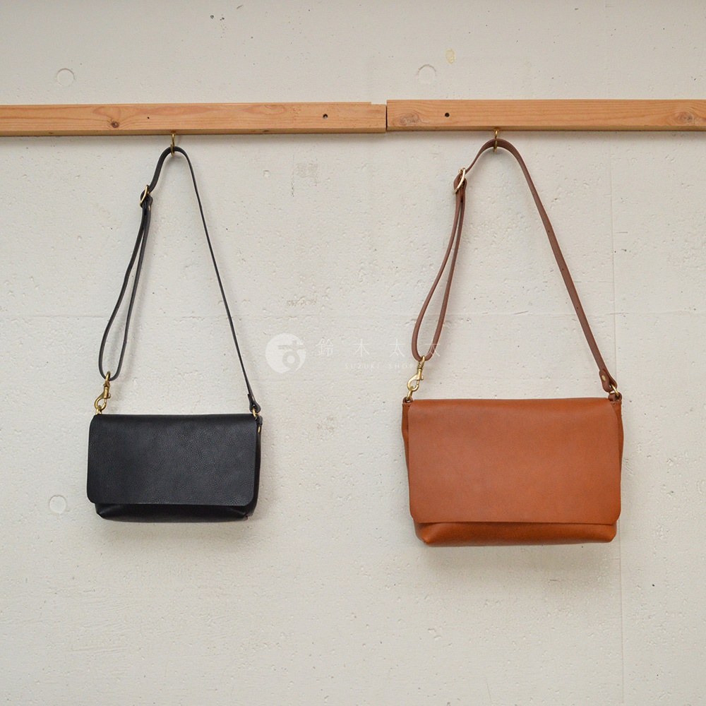 兩個皮革斜背包，一個黑色，一個焦糖色，皆掛於木製橫桿上