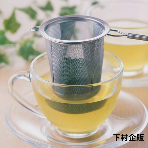 銀色不鏽鋼製深型濾茶網，帶有細緻網孔，置於裝有黃褐色液體的透明玻璃茶杯內，茶杯置於同材質的透明杯碟上，背景有模糊的綠葉裝飾。