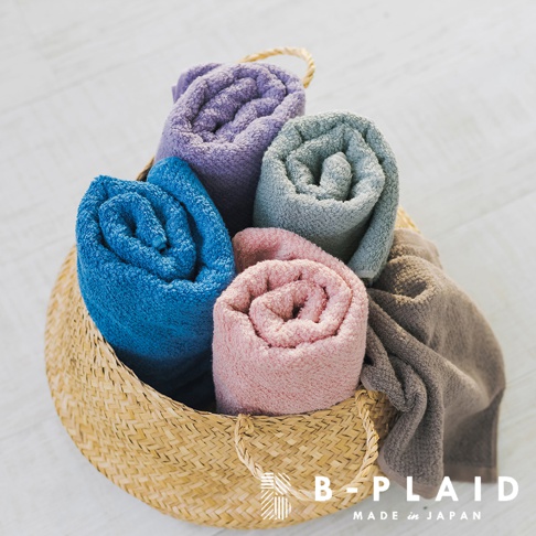 草編籃中盛著五條捲起的毛巾，顏色包含深藍、粉紅、淺灰、紫色和棕褐色。毛巾質地厚實，表面有細緻的紋理。