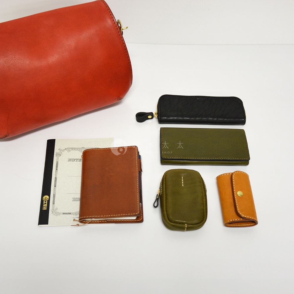紅棕色皮革復古翻蓋郵差包，搭配黑色、綠色、橘色和橄欖綠色皮革小物，包含筆記本、長夾、小零錢包和鑰匙包。
