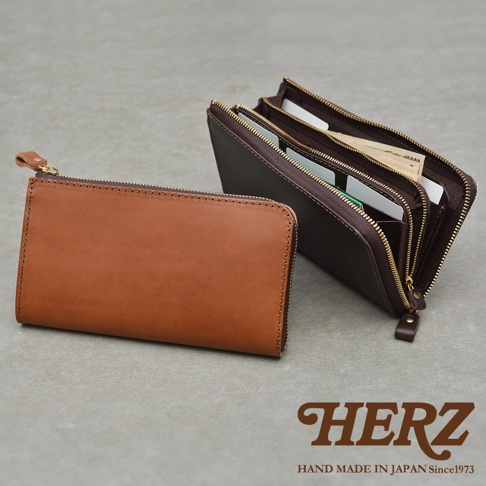 兩個職人手作皮革拉鍊長夾，一個為棕褐色，另一個為深棕色，皆以拉鍊開合，深棕色長夾內部展示著卡片與鈔票，右下角為HERZ品牌標誌與HAND MADE IN JAPAN Since1973字樣。