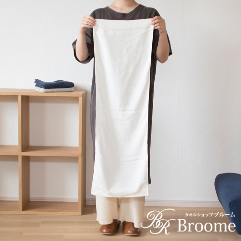 展示一條白色純棉長毛巾，其表面有菱形織紋，頂端有細密的網狀紋路。毛巾由一位身著灰色長洋裝的女士高舉展示。