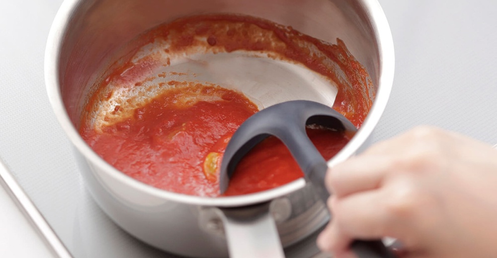 一個沾有番茄醬的銀色鍋子，左手拿著一個灰色的湯勺正在攪拌。