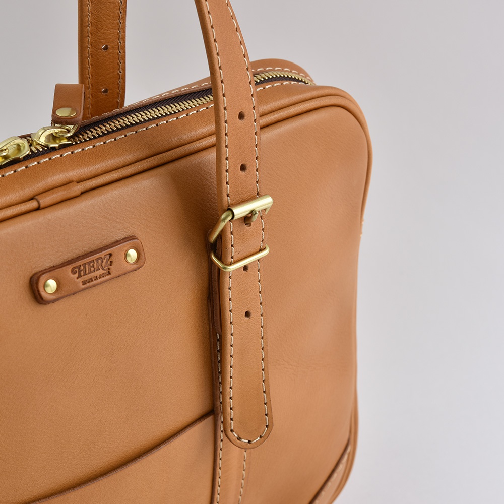 職人手作皮革肩背手提兩用公事包 BW-57，以淺棕色皮革製成，配有金色拉鍊、可調節肩帶和品牌標誌。