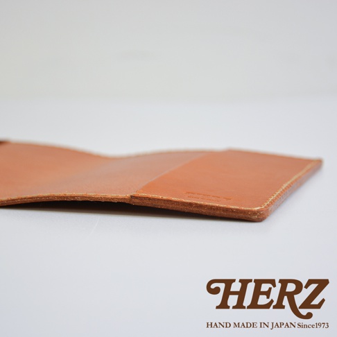一張近距離拍攝的日本HERZ職人手作皮革製品細節圖，展示了厚實棕色植鞣革的邊緣縫線工藝，皮革表面有隱約的品牌壓印，整體呈現溫潤的皮革質感與專業的手工製作細節。