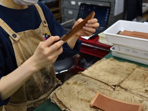 職人正在使用工具加工棕色皮革，桌面上堆疊著未處理的淺棕色皮革材料，背景有白色收納盒和筆記型電腦。