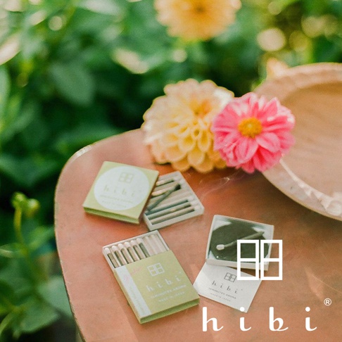 放置於戶外桌面的 hibi 香氛火柴禮盒，柔和的陽光灑落在綠色包裝的火柴盒與一旁盛開的粉色及淺黃色花朵上，盒內展示著細緻的火柴棒與專用燃燒墊，營造出寧靜且優雅的生活香氛氛圍。