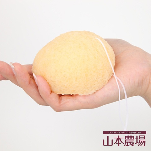 手掌托著一個淡黃色、圓潤的蒟蒻海綿，表面有細密的孔洞，一條白色細繩從海綿側面垂下。