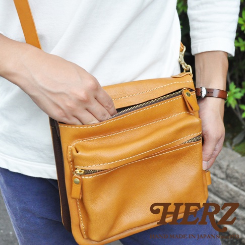 職人手作皮革翻轉雙色直式斜背包，展示為淺棕色皮革，搭配深棕色邊緣，有兩個拉鍊主袋和一個開放式前袋，縫線為白色。