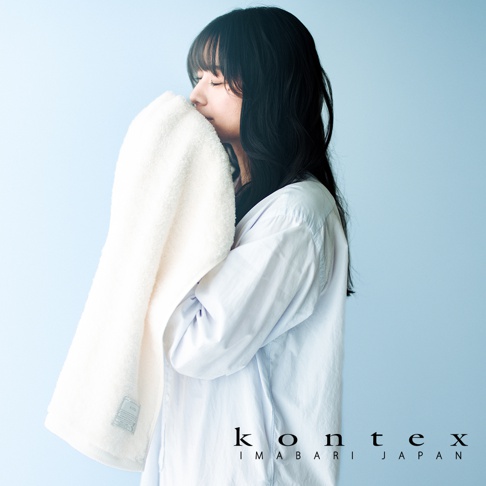 一位留著深色長髮的年輕女性，身穿淺藍色襯衫，雙手捧著一條潔白且質感蓬鬆的 KONTEX 雙效抗菌消臭毛巾輕貼於臉龐，展現出舒適與放鬆的神情，整體構圖簡約柔和，右下方呈現 KONTEX IMABARI JAPAN 品牌字樣。