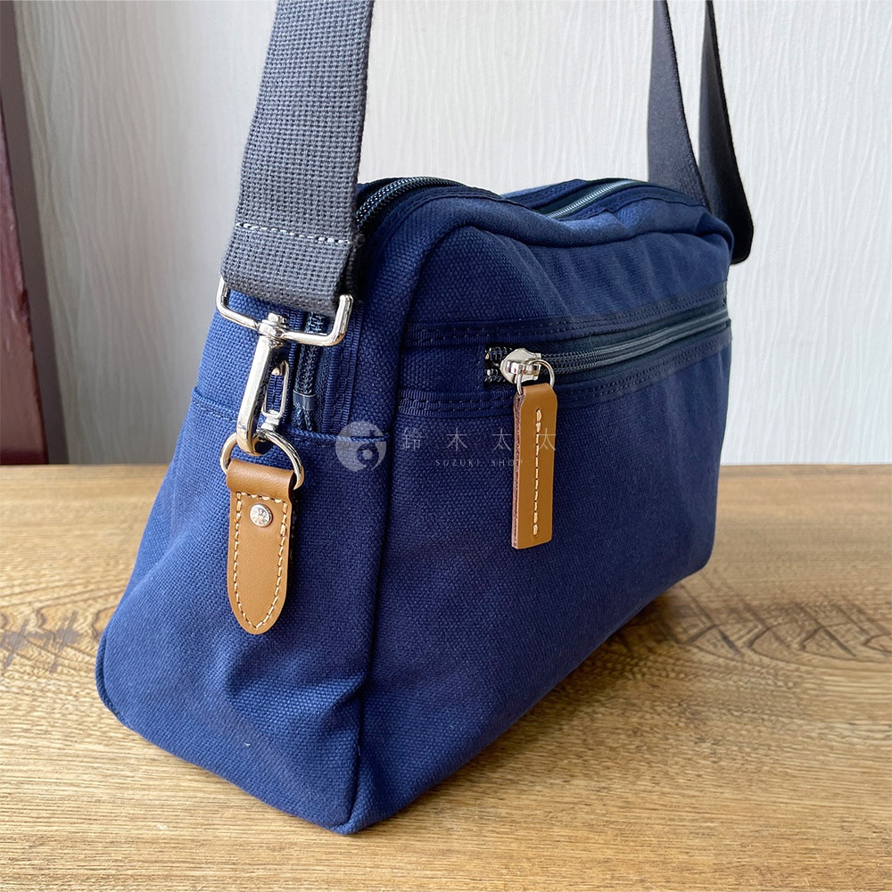 石洗帆布材質的深藍色旅行側背包，配有銀色金屬拉鍊和棕色皮革拉環，肩帶為灰色帆布。