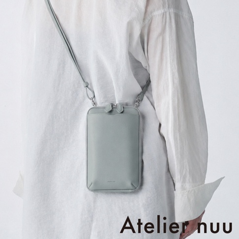 一位身穿白色襯衫的模特兒，側背著一款由 Atelier nuu 設計的 cote 系列鼠尾草綠色皮革隨身手機包，包款呈現簡約俐落的長方形設計，配有精緻的拉鍊細節與同色系背帶，整體風格優雅且現代。