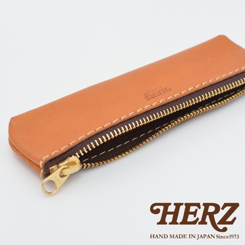 職人手作皮革經典收納筆袋，淺棕色皮革，配有黃銅色拉鍊和清晰的縫線。袋面上印有「HERZ」品牌標誌。