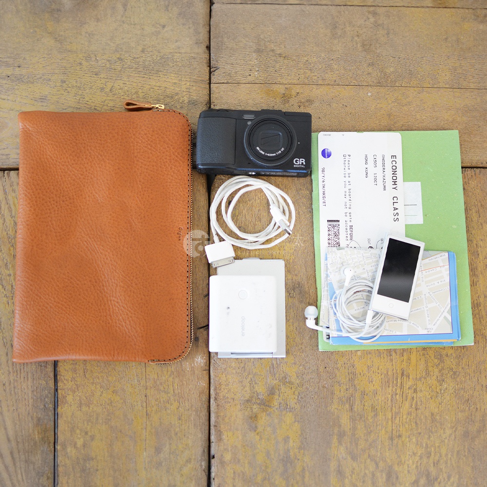 職人手作皮革Organ雙拉鍊收納袋，呈現自然棕色，皮革表面帶有紋理。旁邊擺放著黑色數位相機、白色充電線、白色電源轉接器、綠色機票、白色耳機及 iPod。