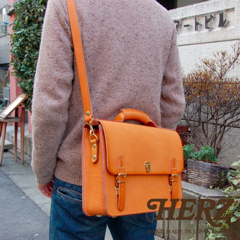 一款橘色皮革手提斜背包，配有金色三扣閉合、頂部提把和可調式肩帶。
