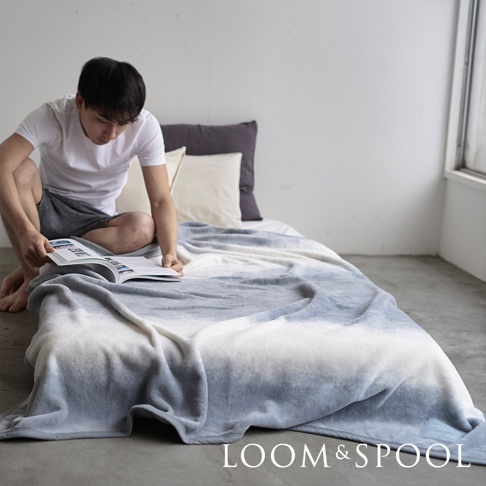 一件藍灰漸層色、短絨毛質地的單人舒柔毯，鋪在床上，一名男子坐在床邊閱讀。