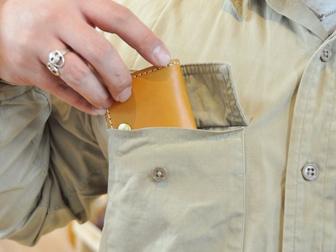 淺棕色皮革箱型零錢包，邊緣有白色縫線，置於卡其色襯衫的胸前口袋中，一只戴著銀色笑臉戒指的手正在將零錢包放入口袋。