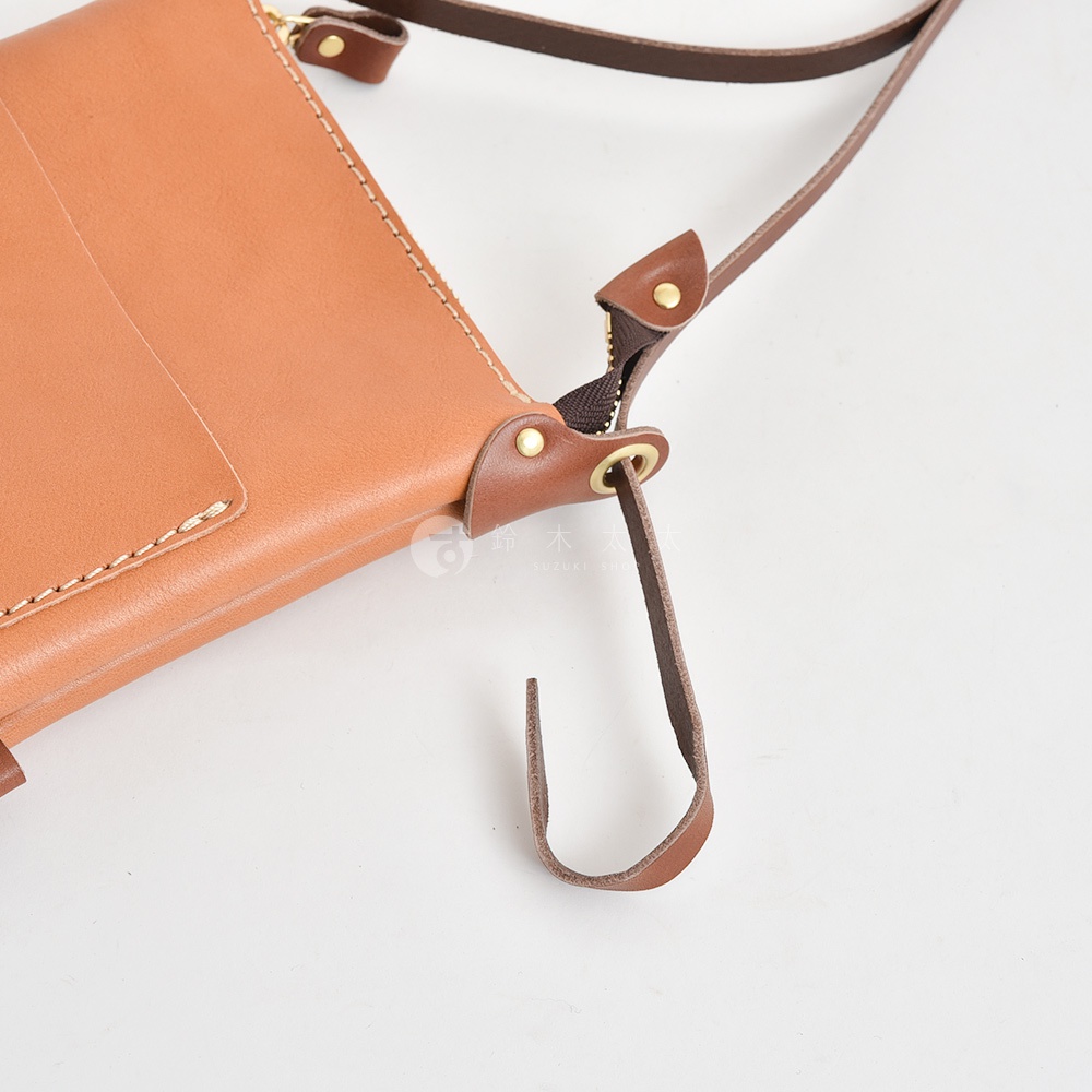 淺棕色皮革直式隨身單肩包，帶有深棕色皮革肩帶、拉鍊和飾邊。包包邊緣有白色縫線，並有一個金屬環。