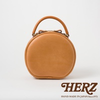 HERZ 品牌職人手作皮革兩用圓形包 Q-13，採用經典棕褐色皮革，搭配簡約提把與品牌 Logo，展現日式職人精神。