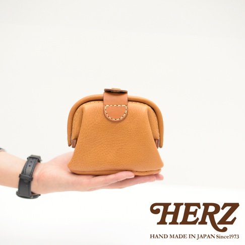 職人手作皮革口金化妝包，淺棕色，皮革材質，帶有閉合扣，由手托起，背景純白。