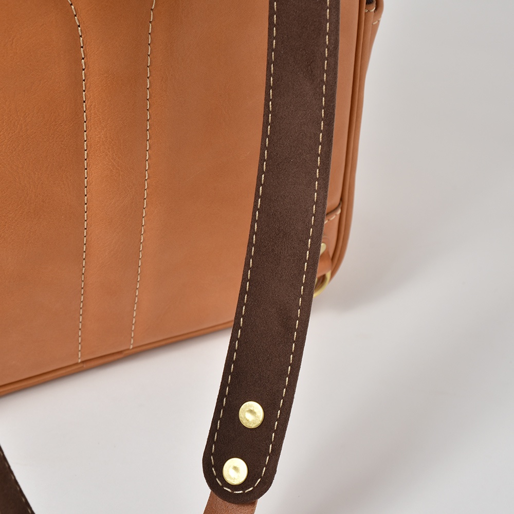 職人手作皮革多功能後背包 R-21，展現光滑棕色皮革主體，搭配深棕色麂皮肩帶，以白色縫線勾勒出細緻結構，並以黃銅色金屬鉚釘固定。