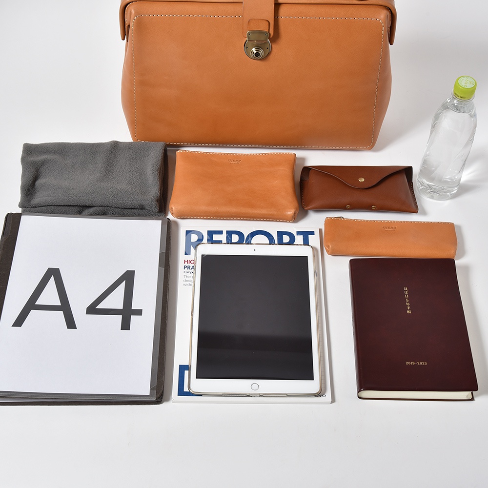 職人手作皮革軟式杜勒斯三用背包 BJ-16，展示於圖片中，背包為淺棕色皮革材質，設有金色金屬鎖扣。周圍擺放有灰色毛巾、淺棕色皮革化妝包、棕色皮革眼鏡盒、淺棕色筆袋、A4文件夾、雜誌、平板電腦和一本深棕色筆記本。