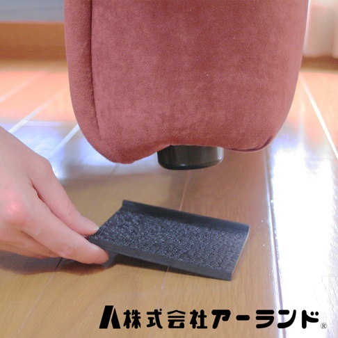 一塊深灰色、表面帶有顆粒紋理的沙發防滑墊，置於木地板上，一隻手正將其放置在紅色絨面沙發腳下方。