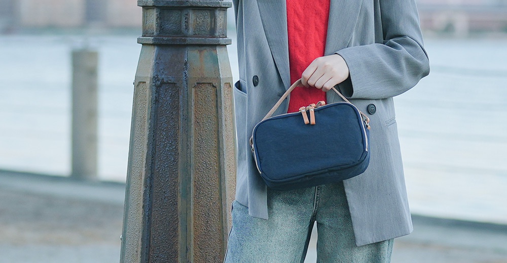 一位身穿灰色西裝外套和紅色上衣的女性，肩上斜背著一個深藍色的方形包包，包包帶為米白色。