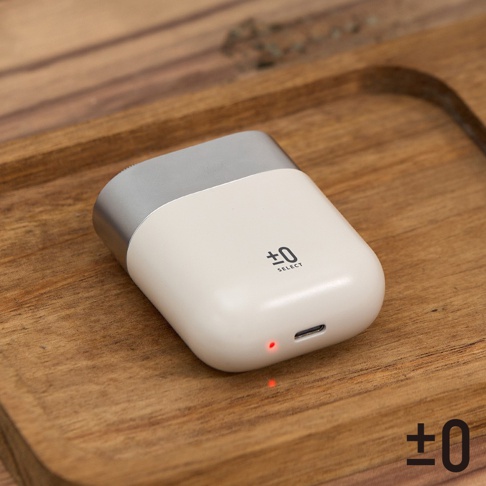 象牙白與銀色組合的便攜式刮鬍刀，表面印有「±0 SELECT」標誌，機身底部設有 USB-C 充電孔，紅色的指示燈亮起，產品置於木紋表面。