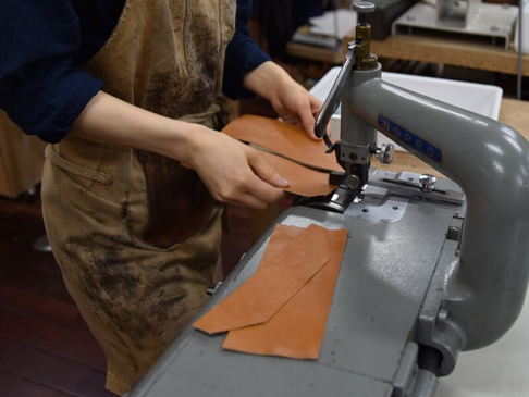 職人身穿帆布圍裙，手持棕褐色皮革，於縫紉機上製作簡約輕薄扁長夾。