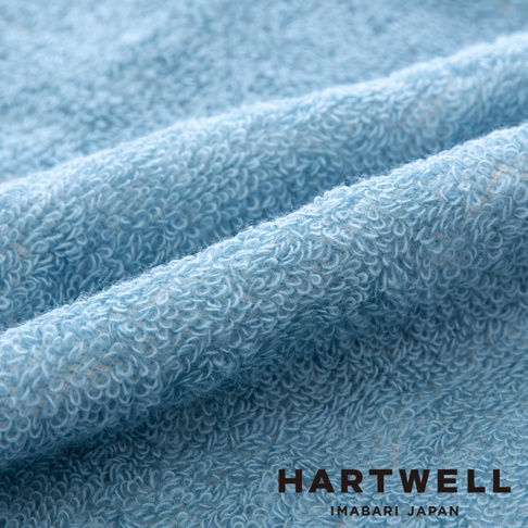 淺藍色毛巾布料，質地柔軟，紋理清晰，表面有細緻的毛圈，布料呈現自然的皺褶，印有 HARTWELL IMABARI JAPAN 字樣。