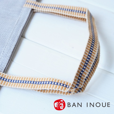 灰色布質提把蔬果收納袋，提把織帶為米白色，帶有藍色與黃褐色直條紋，質料為棉麻。