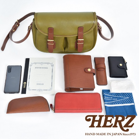 橄欖綠皮革傳統釣魚包，搭配棕色皮革翻蓋與束帶，附有金屬扣。周圍擺放著棕色皮夾、紅色長夾、黑色小錢包、筆記本、手機、手機套及印有藍白圖案的布巾。