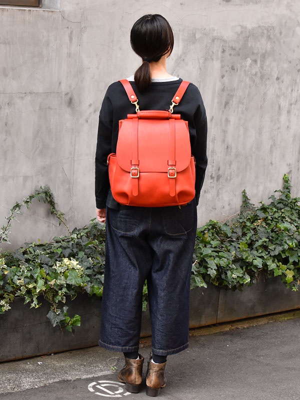 紅色皮革雙扣後背包，有頂部提把和可調節的肩帶，搭配金色金屬扣環。