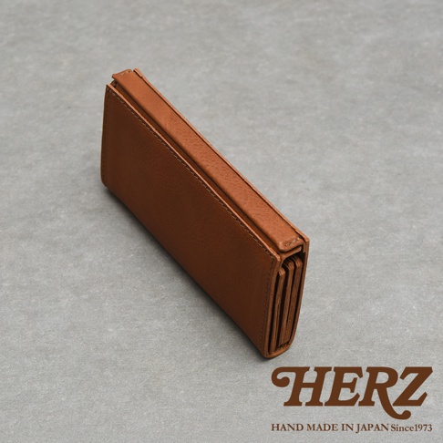 從高處視角拍攝的一個日本 HERZ 職人手作皮革長夾，材質呈現溫潤的棕色皮革質感，外觀簡約具有獨特的風琴摺疊結構，右下角印有品牌名稱與手工製作資訊，整體設計充滿日式職人匠心風格。