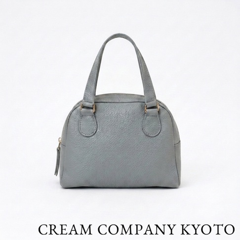 CREAM COMPANY 京都出品的灰色手作半月迷你手提包，採用柔軟的glove牛皮革材質製作，包身紋理細緻且外觀簡約大方，底部標註有品牌名稱，展現精緻的職人手作工藝。