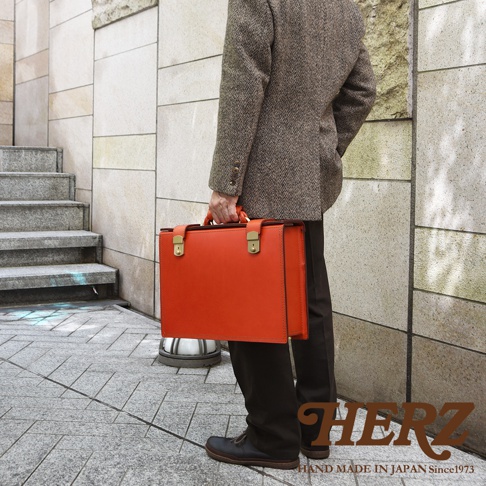 職人手作皮革機師箱 BC-31，亮橘色皮革，配有金色鎖扣和提把。箱體方正，線條簡潔，展現經典設計。