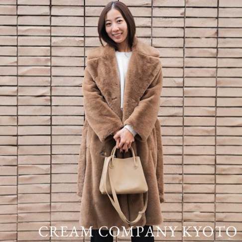 身穿棕色長款毛絨外套的女性，手持米色皮革兩用手提托特包，包款簡潔，搭配可拆卸肩帶。