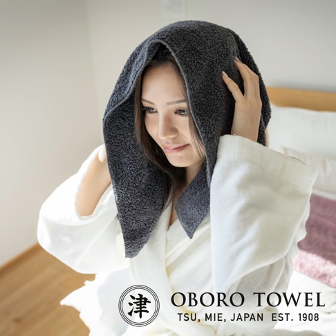 一位女子頭上戴著深灰色、紋理柔軟的擦髮巾，身穿白色浴袍，手調整著擦髮巾，臉部朝向右側。