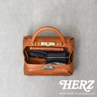 職人手作皮革Organ兩用口金手提包，採用天然皮革製成，呈現溫潤的焦糖棕色。包款設計簡潔，有手提把和金屬鎖扣，內部可見皮夾、零錢包等物品，展現其收納功能。