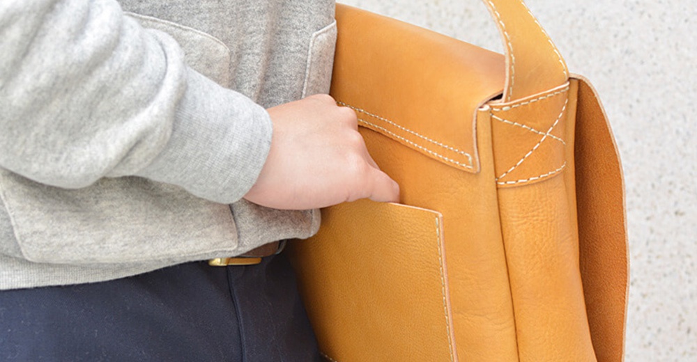 一個人的手從側面靠近一個黃褐色的皮革斜背包，包包上有著縫線細節。