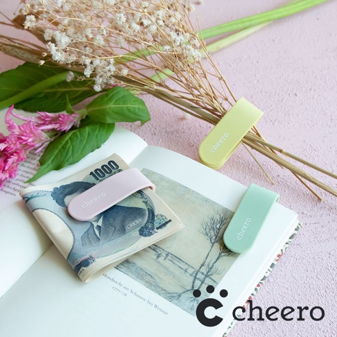 粉色、淺綠色和黃色的萬能QQ變形夾，夾在日圓紙鈔和書本上，背景為乾燥花束與粉色紋理桌面。