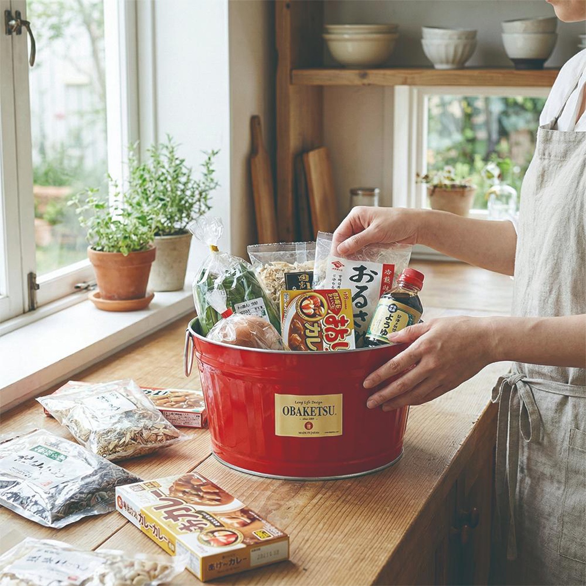 一個裝滿雜貨的紅色OBAKETSU金屬桶，放在廚房的木製檯面上，背景是窗戶和架子。