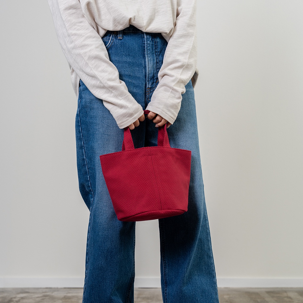 紅色三河木綿刺子織圓底水桶包，配有雙提把，由一位身穿淺色長袖上衣和藍色牛仔褲的人士手持。