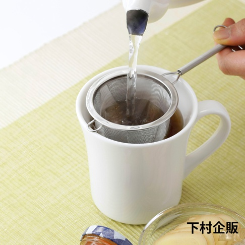 不鏽鋼深型濾茶網，網目細緻，帶有可掛於杯緣的掛環和彈簧手柄，正置於白色馬克杯中，熱水正從茶壺注入，濾網內有茶葉。