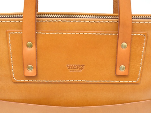 職人手作皮革威靈頓直式拉鍊托特包 (T-151) 的特寫。包身為淺棕色皮革，飾有同色系皮革提把及金色卯釘。正面有壓印的「HERZ」品牌標誌與「MADE IN JAPAN」字樣，周圍以白色縫線勾勒出精緻的口袋輪廓。