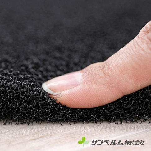 特寫鏡頭展示 Sanbelm 品牌的浴墊用透氣防滑墊，一位成人的手指正輕壓在黑色、具有多孔隙結構的立體網狀材質表面上，強調其優異的透氣性與彈性，畫面右下方印有品牌名稱與標誌。