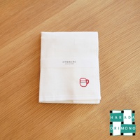 白色八重紗材質擦拭巾，表面有網格紋理，左下角繡有紅色馬克杯圖案，中央有白色紙帶，印有日文與英文產品資訊。